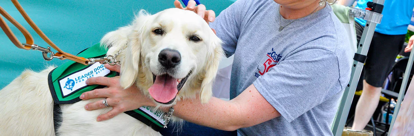 A cream-colored dog wearing a green Leader Dogs for the Blind vest is happily looking at the camera. A woman is next to the dog, with one hand on his chest and one hand petting the dog's head.