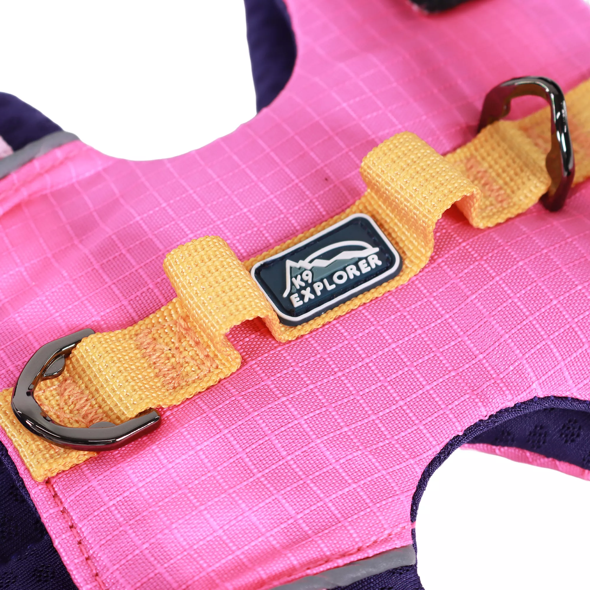 A close‑up view of the Rosewood‑colored full‑coverage dog harness is shown against a white background. The harness features a bright pink fabric panel with a grid‑stitched pattern. Across the top of the panel is an orange woven strap that includes two metal D‑rings positioned at each end. Between the D‑rings are two orange fabric loops stitched into the strap for attaching accessories. A small woven brand patch with mountain graphics is sewn onto the center of the orange strap.